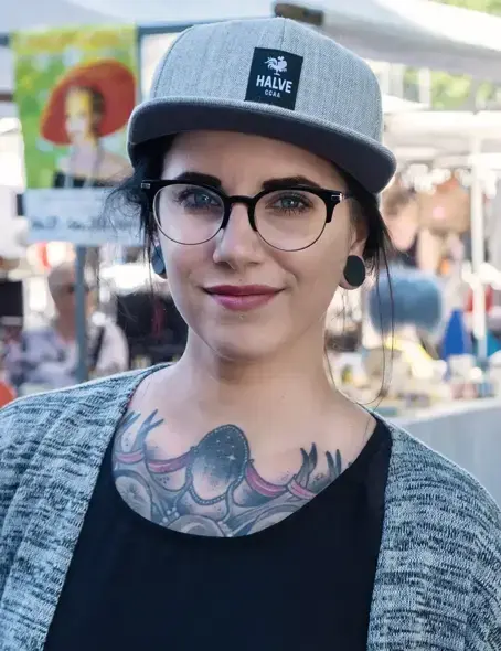 Die 33-j&auml;hrige Nicci blickt grinsend in die Kamera. Sie tr&auml;gt eine Brille, eine Kappe und ein dunkles Shirt, worunter Tattoos zu erkennen sind.