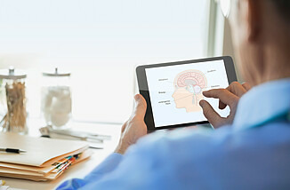 Ein Arzt betrachtet die grafische Ansicht eines Gehirns auf einem Tablet. Vor ihm auf dem Tisch liegen Akten. 