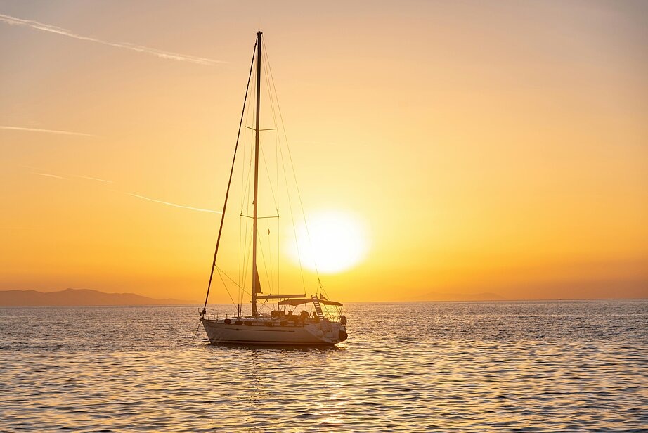 Ein Segelboot ohne gehisste Segel fährt auf ruhiger See vor dem Sonnenuntergang auf dem Meer, im Hintergrund sind verschwommene Bergformationen zu sehen.