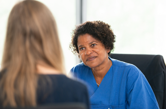 Dr. Geraldine de Heer ist in ein Gespräch mit einer Patientin vertieft. Beide sitzen sich gegenüber. Dr. Geraldine de Heer ist von vorne zu sehen, die Patientin von hinten.