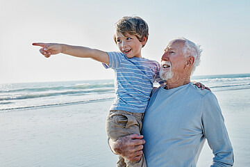 Ein älterer Mann steht am Strand. Er trägt einen Jungen auf dem Arm, der in die Ferne zeigt. Beide schauen in die gleiche Richtung und sehen zuversichtlich aus.