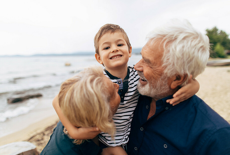 Ein älteres Paar, ein Mann und eine Frau, sind am Strand. Sie tragen einen kleinen Jungen in ihrer Mitte auf den Armen und umarmen sich dabei. Alle drei lachen.