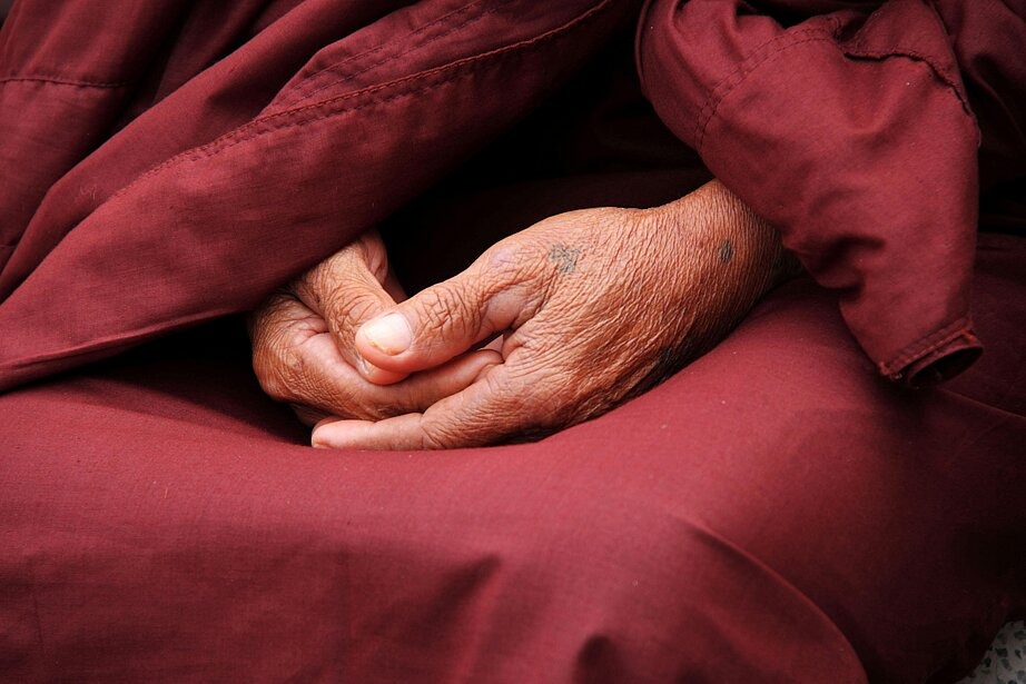 Zu sehen sind die im Schoß eines buddhistischen Mönches zusammengelegten, faltigen Hände in Nahaufnahme. Er trägt eine dunkelrote Robe.