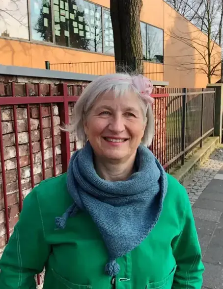 Die 58-j&auml;hrige Elke steht drau&szlig;en auf dem Gehweg und l&auml;chelt. Sie hat graue Haare, die etwas &uuml;ber ihre Ohren gehen, und tr&auml;gt einen rosa Haarreif. Sie hat eine gr&uuml;ne Jacke und einen blauen Schal an.