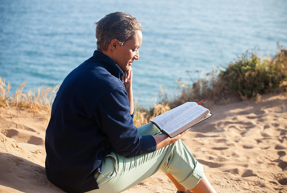 Eine junge Frau sitzt am Strand und liest in der Bibel. 