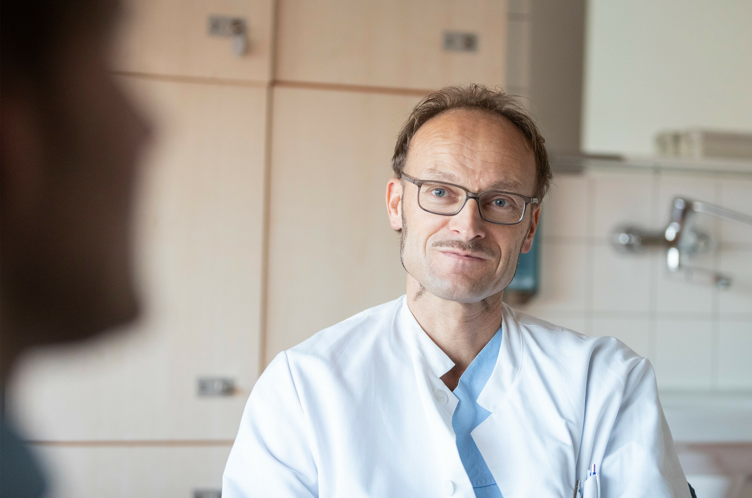 Dr. Frank Logemann im Gespräch mit einem Patienten, der im Vordergrund nur als Umriss erkennbar ist. Herr Dr. Logemann hört aufmerksam zu.