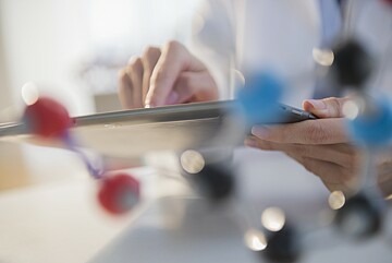 Die Hand eines Arztes ist im Hintergrund zu erkennen, wie sie auf ein Tablet tippt. Davor ist verschwommen ein Molekülmodell zu sehen. 