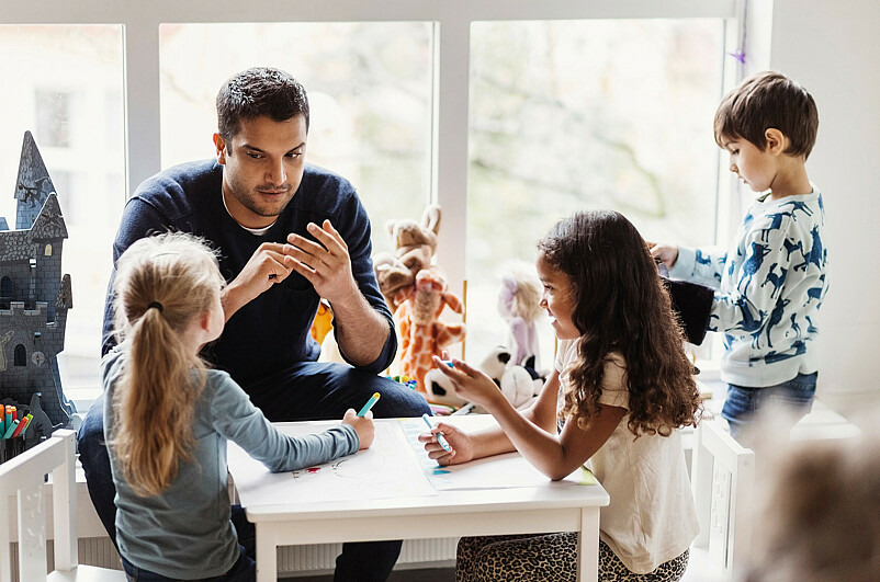 Ein Mann sitzt an einem Tisch mit einer Gruppe Kinder und erklärt ihnen etwas.