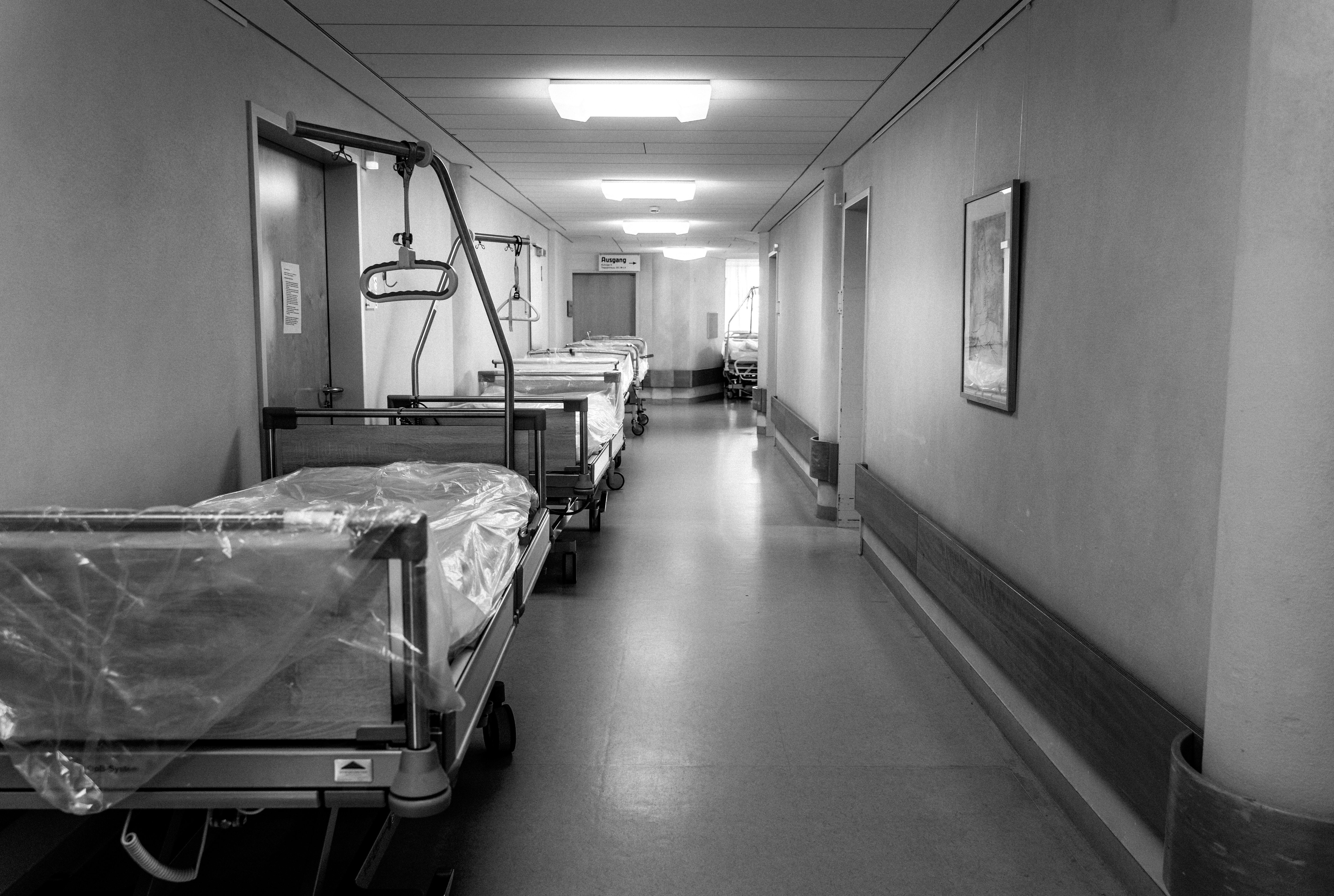 Das Foto zeigt einen Krankenhausflur. Auf der linken Seite des Flures stehen zwei mit Folie bedeckte Krankenhausbetten hintereinander. Die Farben des Fotos sind gräulich.