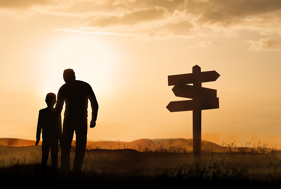 Zu sehen ist ein Vater mit seinem Sohn, die im Sonnenuntergang stehen und von hinten fotografiert wurden. Man sieht lediglich ihre Umrisse, während rechts von ihnen ein schwarzes Schild steht, dessen Wegweiser in verschiedene Himmelsrichtungen zeigen.