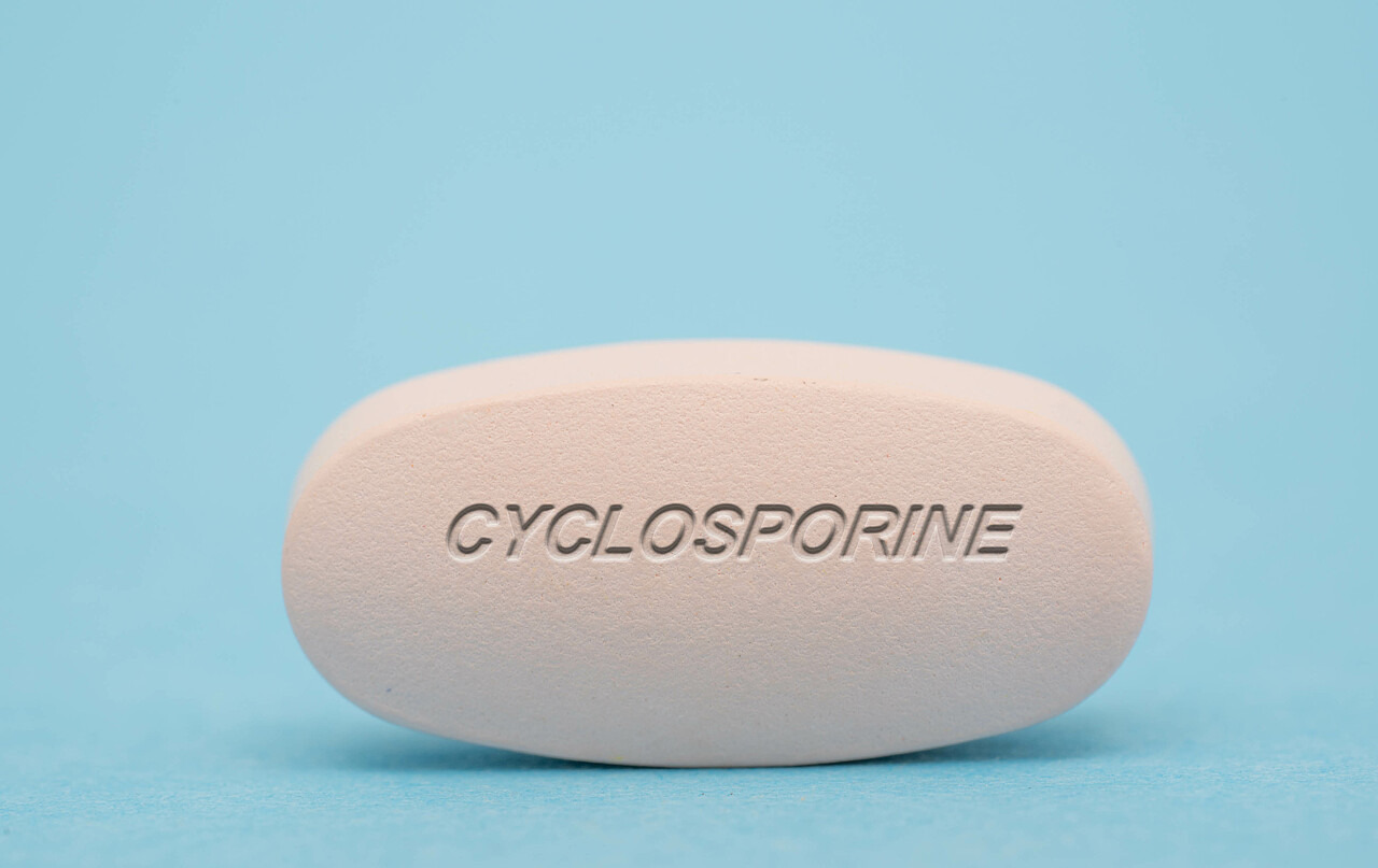 Zu sehen ist eine Nahaufnahme einer weißen Tablette. Auf ihr steht „Cyclosporine“. Sie liegt auf einer hellblauen Fläche.
