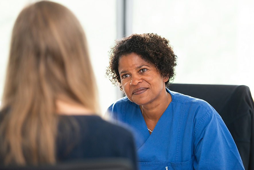 Dr. Geraldine de Heer ist in ein Gespräch mit einer Patientin vertieft. Beide sitzen sich gegenüber. Dr. Geraldine de Heer ist von vorne zu sehen, die Patientin von hinten.