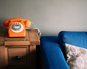 Ein orangenes Wählscheibentelefon steht auf einem Holztisch. Daneben steht ein blaues Sofa.